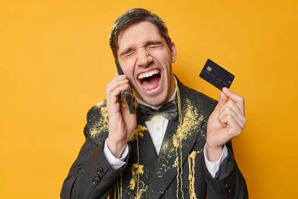 Man in tuxedo and covered in party paraphernalia against mustard yellow background yells in joy into cell phone while holding a credit card in his other hand.